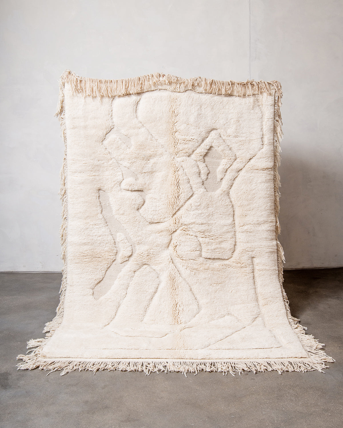 Berberlin x Malaika Raiss Beni M'rirt rug made of velvety cream sheep's wool with natural wool tassels on all four sides, being held up against a white wall and concrete floor.